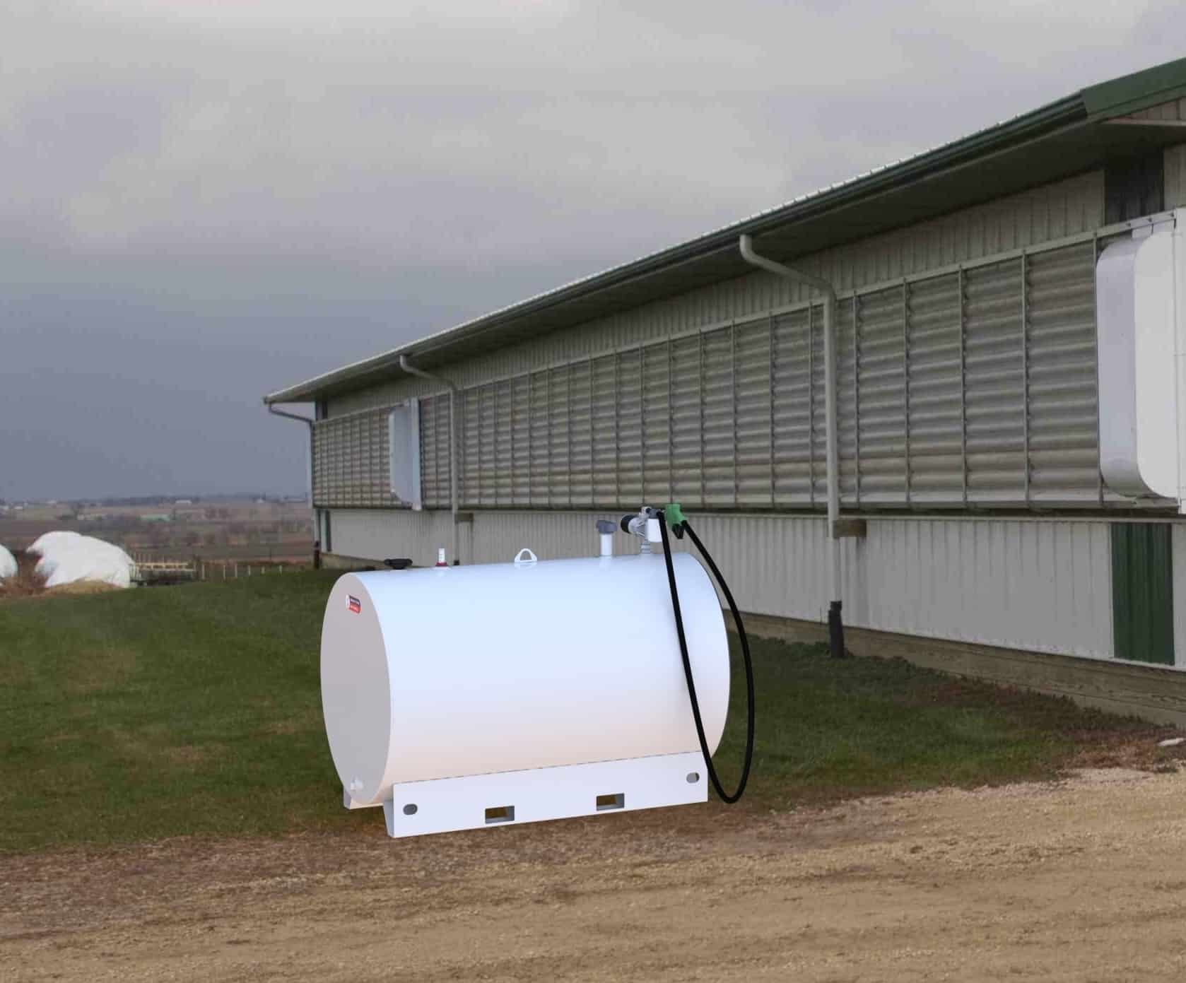White horizontal fuel storage tank with a black hose and green fuel nozzle mounted on a metal skid, positioned on grass beside a large agricultural building with metal siding.